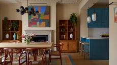 a kitchen come dining area with a colorful piece of art above the mantelpiece in front of the dining table with a black pendant light hanging above and blue kitchen cabinetry and wooden cabinets either side of the fireplace filled with decor and plants