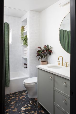 white and green bathroom with terrazzo tiled floor, gray vanity, gold taps, green shower curtain, built in storage, white wall tiles, plants