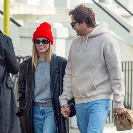 Margot Robbie walks down the street in Notting Hill, London, wearing cropped jeans with Converse and a grey knit with a long-line leather jacket and red beanie.