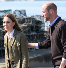 Prince William wearing a brown sweater, putting his hand on Kate Middleton's back as she walks in front of him in a green blazer and matching sweater