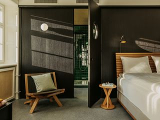 A room in the hotel. Gray floors, with a wooden bed to the right, and a chair to the left. Dark brown doors are open, and we see the bathroom shower that has dark green tiles.