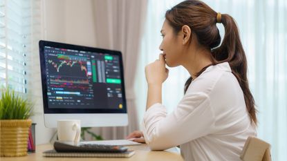 A young woman looks at her computer screen, which is filled with stock charts and other graphics. 