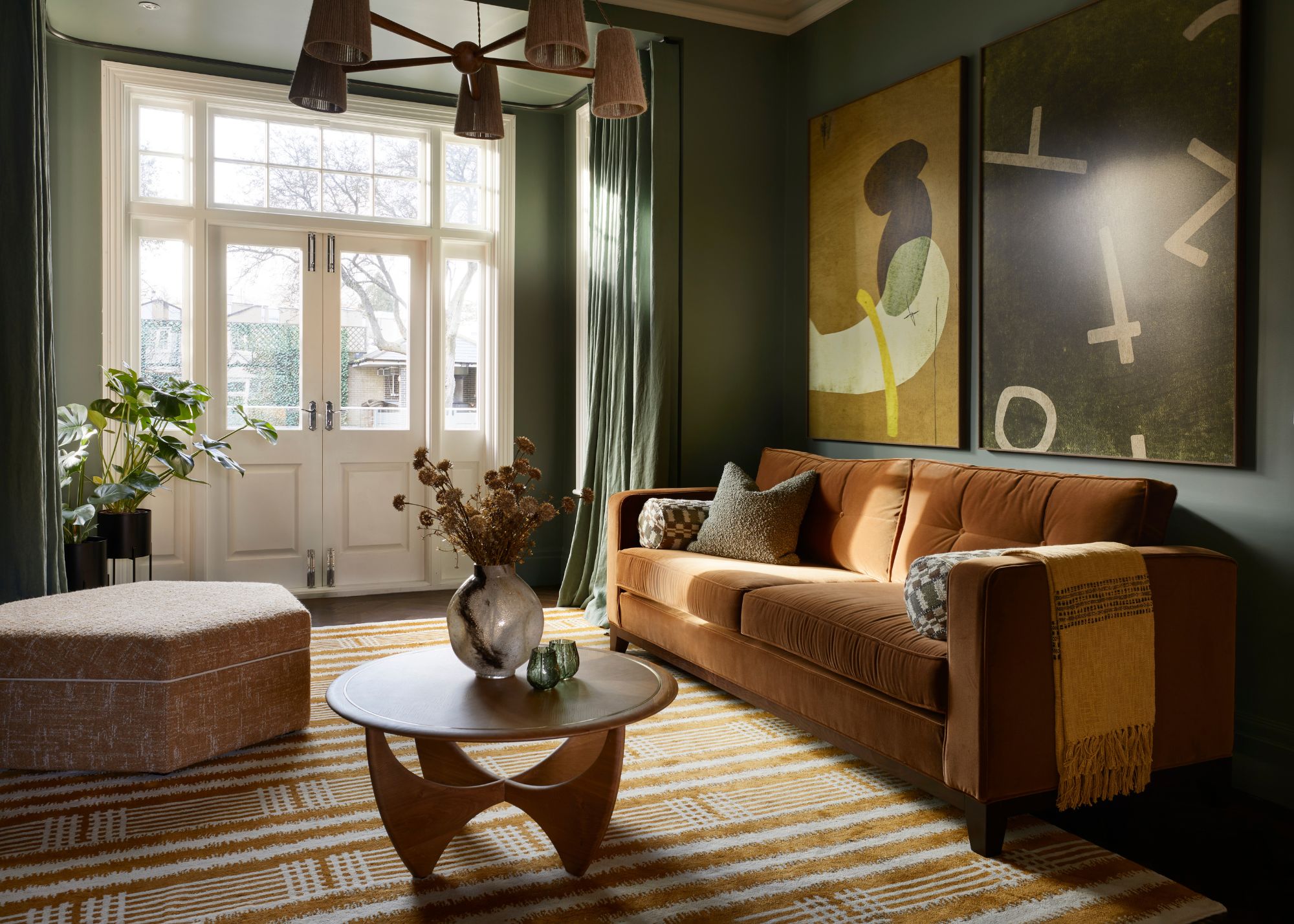 A cozy living room with dark green walls, French doors leading outside, green curtains, a burnt orange sofa with an ochre and cream modern patterned rug, a round wood coffee table, and two dark yellow and green abstract artworks on the wall.