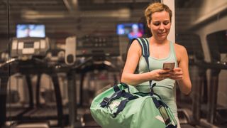 Woman looks at her phone with gym in background, a gym bag is slung over her shoulder biceps curl with dumbbells