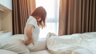 woman with long hair sits up in bed with her back hunched and hands at her waist, as if in pain