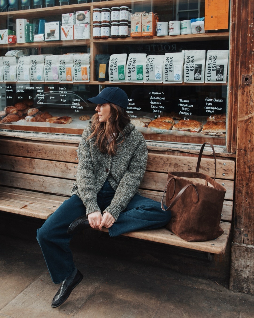 Woman wears cap, green jumper, blue jeans. loafers and suede bag