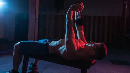 Muscular man exercise with dumbbell lieas on bench in red-blue gradient light in dark gym hall