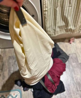 A cream linen bag with black handles full of laundry being emptied onto a gray linoleum kitchen floor next to the washer