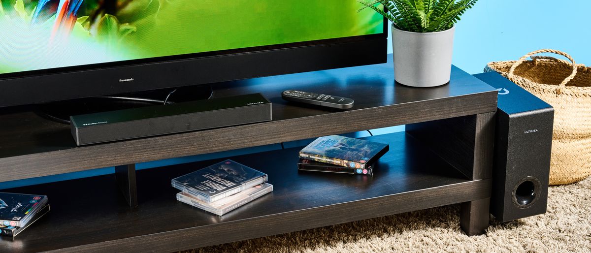 the ultimea nova s50 a black compact soundbar with LED on the front and a wired subwoofer is photographed on a black TV mount connected to a panasonic TV 