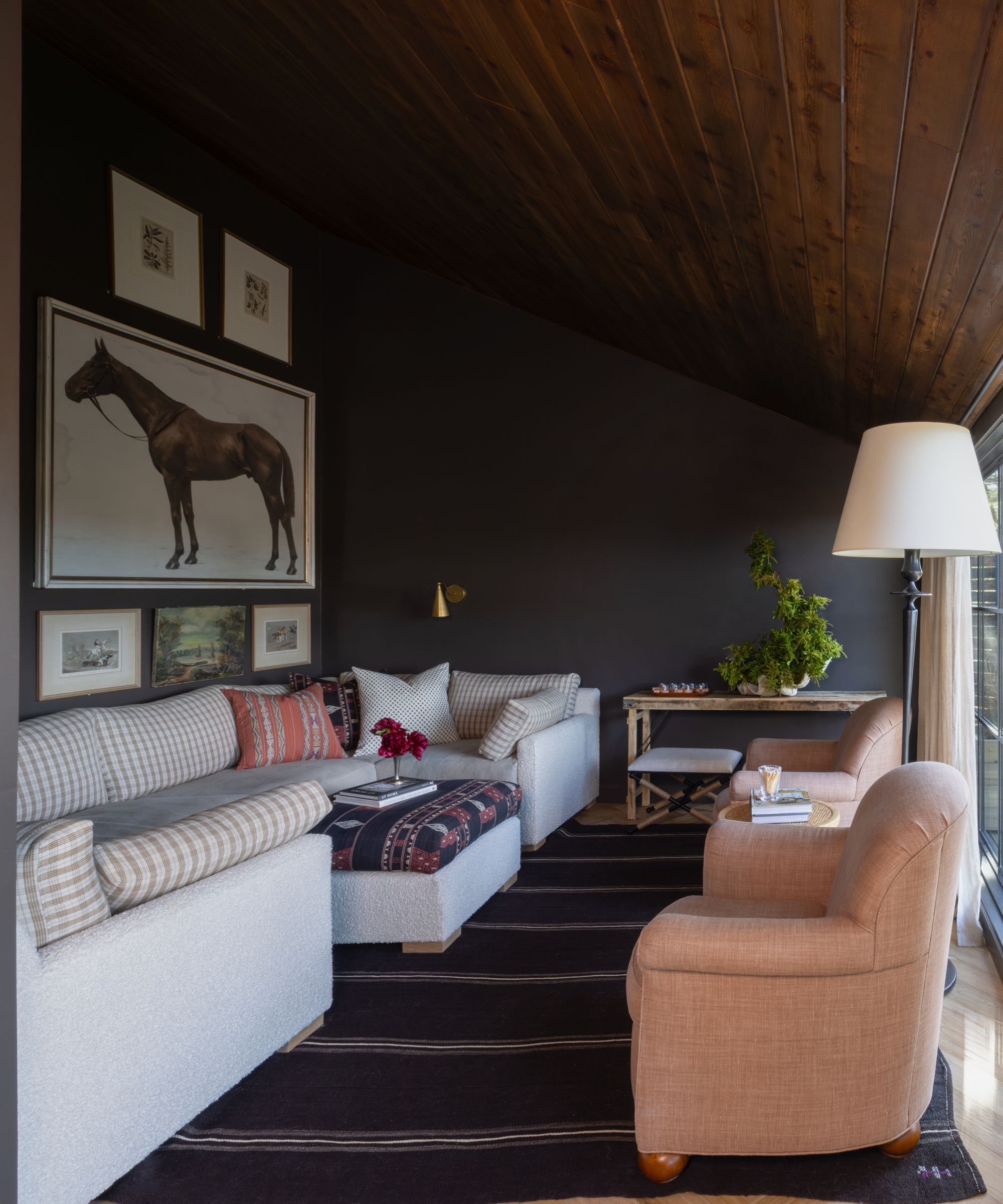 A dark snug with soft seating, including a gingham upholstered sectional and peach armchairs. The dark walls feature framed equestrian artwork and there is a dark striped rug on the floor