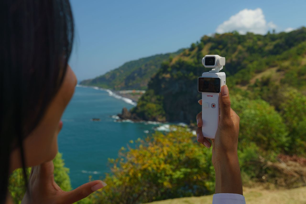 HoverAir Versa 2-in-1 drone and pocket gimbal camera in white, as a gimbal camera in person's hand by a dramatic coastline on a sunny day