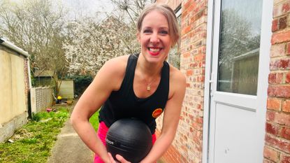Woman stands on garden path next to a house holding a medicine ball