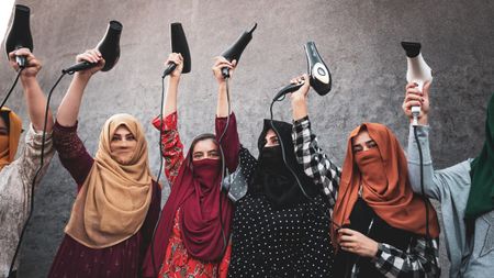 A row of Afghan refugee women in brightly coloured headscarves, most with their faces veiled, stand against a grey concrete wall holding hairdryers triumphantly above their heads, cables dangling. 