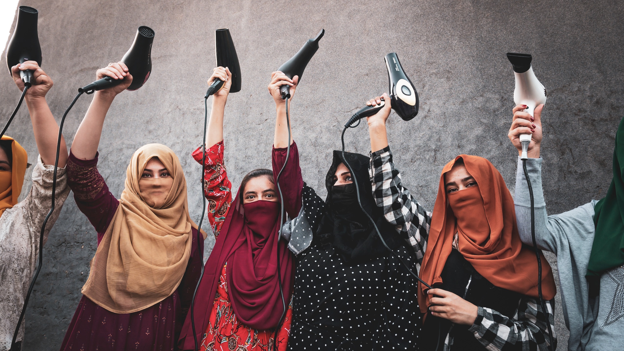 A row of Afghan refugee women in brightly coloured headscarves, most with their faces veiled, stand against a grey concrete wall holding hairdryers triumphantly above their heads, cables dangling. 