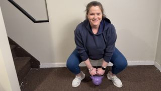 Trainer and fitness writer Jennifer Rizzuto squats in her hallways holding a kettlebell. Her arms are straight and the kettlebell is dangling towards the ground as she smiles at the camera.