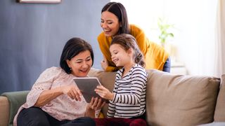 Grandmother, mother and granddaughter sitting on sofa at home, using a tablet