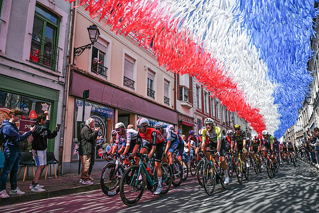 TOPSHOT - The pack of riders (peloton) cycles under decorative streamers hung above the race route in Aire-sur-la-Lys during the 3rd stage of the 112th edition of the Tour de France cycling race, 178.3 km between Valenciennes and Dunkerque (Dunkirk), Northern France, on July 7, 2025. (Photo by Loic VENANCE / AFP) (Photo by LOIC VENANCE/AFP via Getty Images)