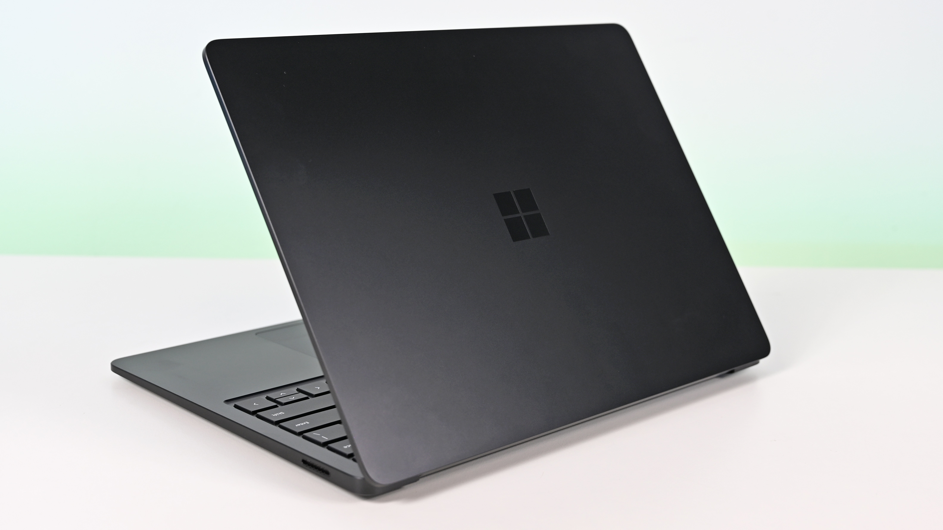 Surface Laptop for Business 8th Edition with a closed screen featuring a Windows logo, resting on a white surface. The background is a soft gradient from light blue to green.