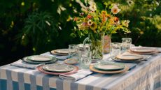 A garden tablescape with a striped tablecloth, stacked plates, clear glasses and a vase of colorful flowers