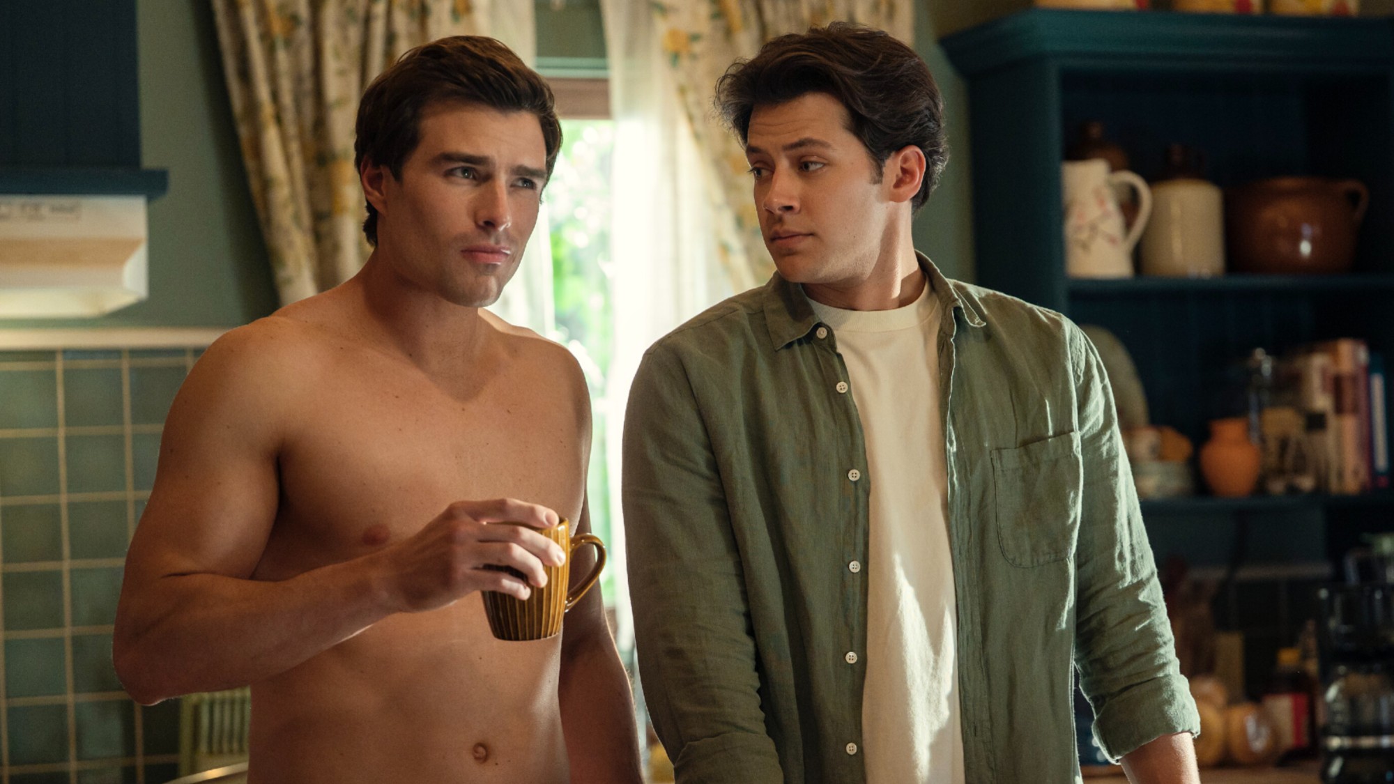 From left to right: Charlie standing without a shirt on and holding a cup of coffee and Sam standing next to him, in a shirt.