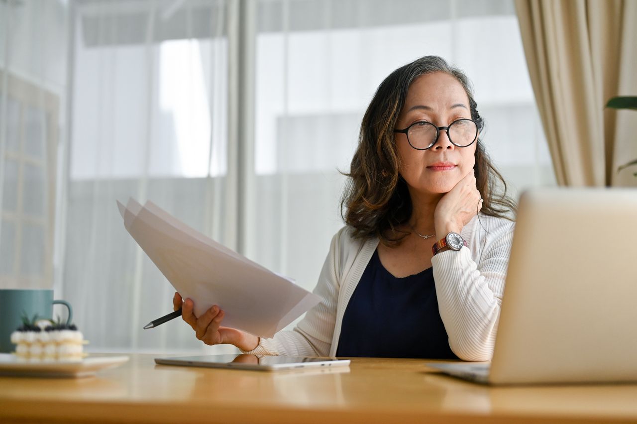 Concentrating Asian middle aged female teacher or businesswoman in glasses sitting at desk using portable computer and examining paperwork. Age and technology