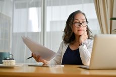 Concentrating Asian middle aged female teacher or businesswoman in glasses sitting at desk using portable computer and examining paperwork. Age and technology