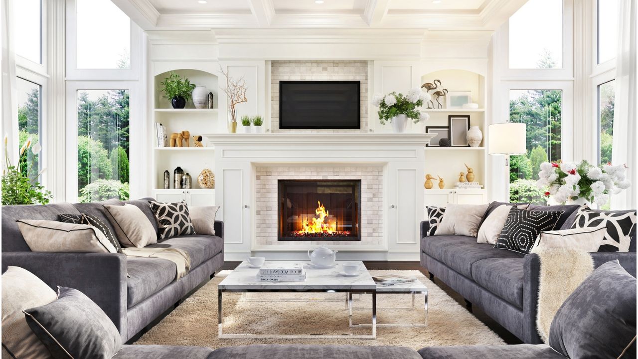 Large white bright living room with floor to ceiling windows either side of a white tall chimney stack with a roaring fire in daylight