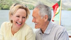 An older couple laugh with each other while on a boat, a flag for Portugal in the background. 