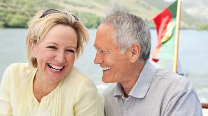 An older couple laugh with each other while on a boat, a flag for Portugal in the background. 