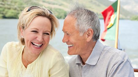 An older couple laugh with each other while on a boat, a flag for Portugal in the background. 