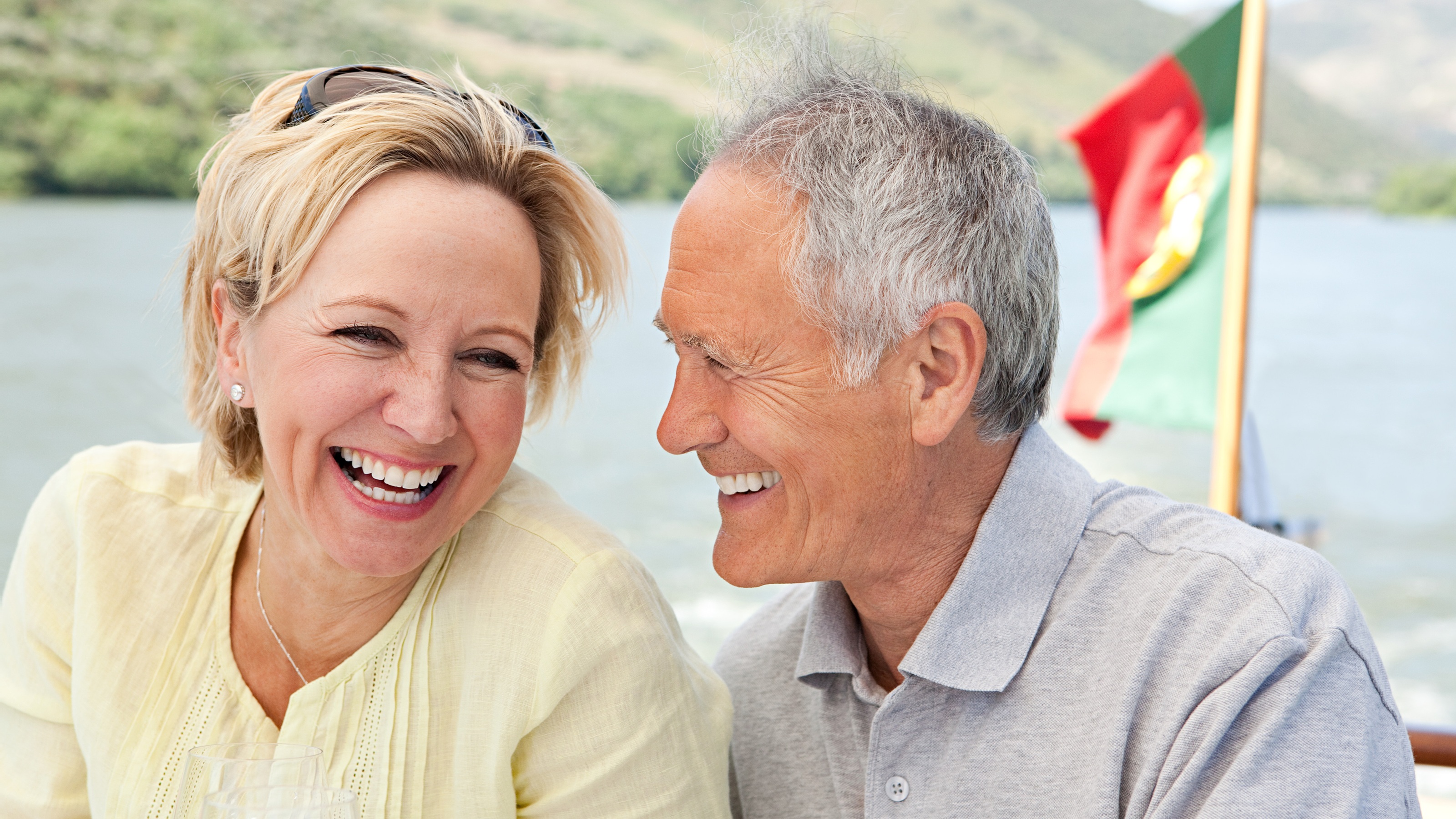An older couple laugh with each other while on a boat, a flag for Portugal in the background. 