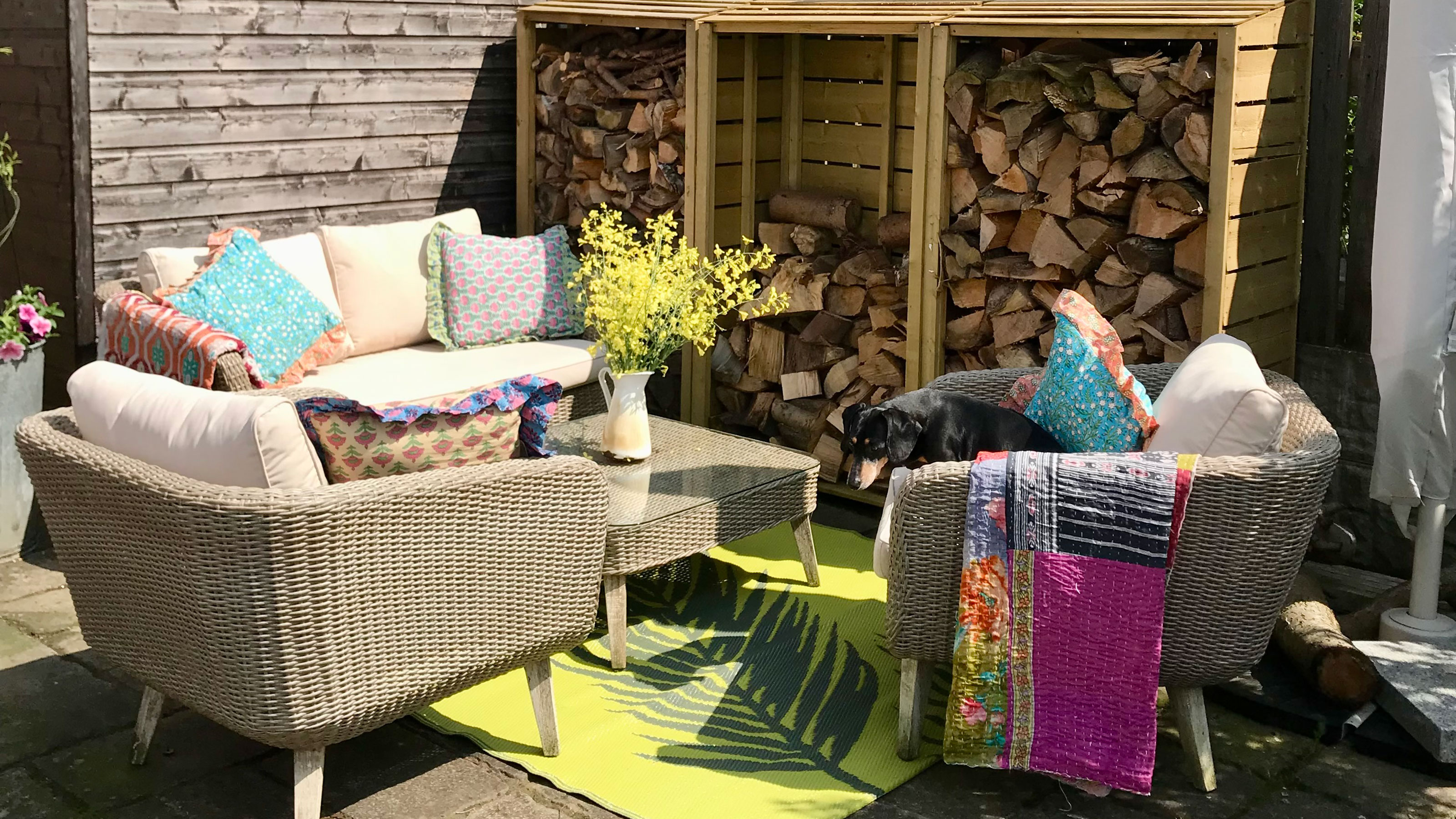 Outdoor sofa and armchairs with colourful cushions and throws around coffee table on top of bright coloured rug