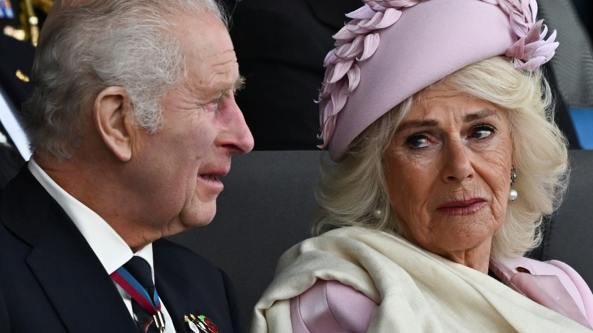 Queen Camilla wears a pink hat and pink suit jacket with a cream scarf and looks tearfully at husband King Charles