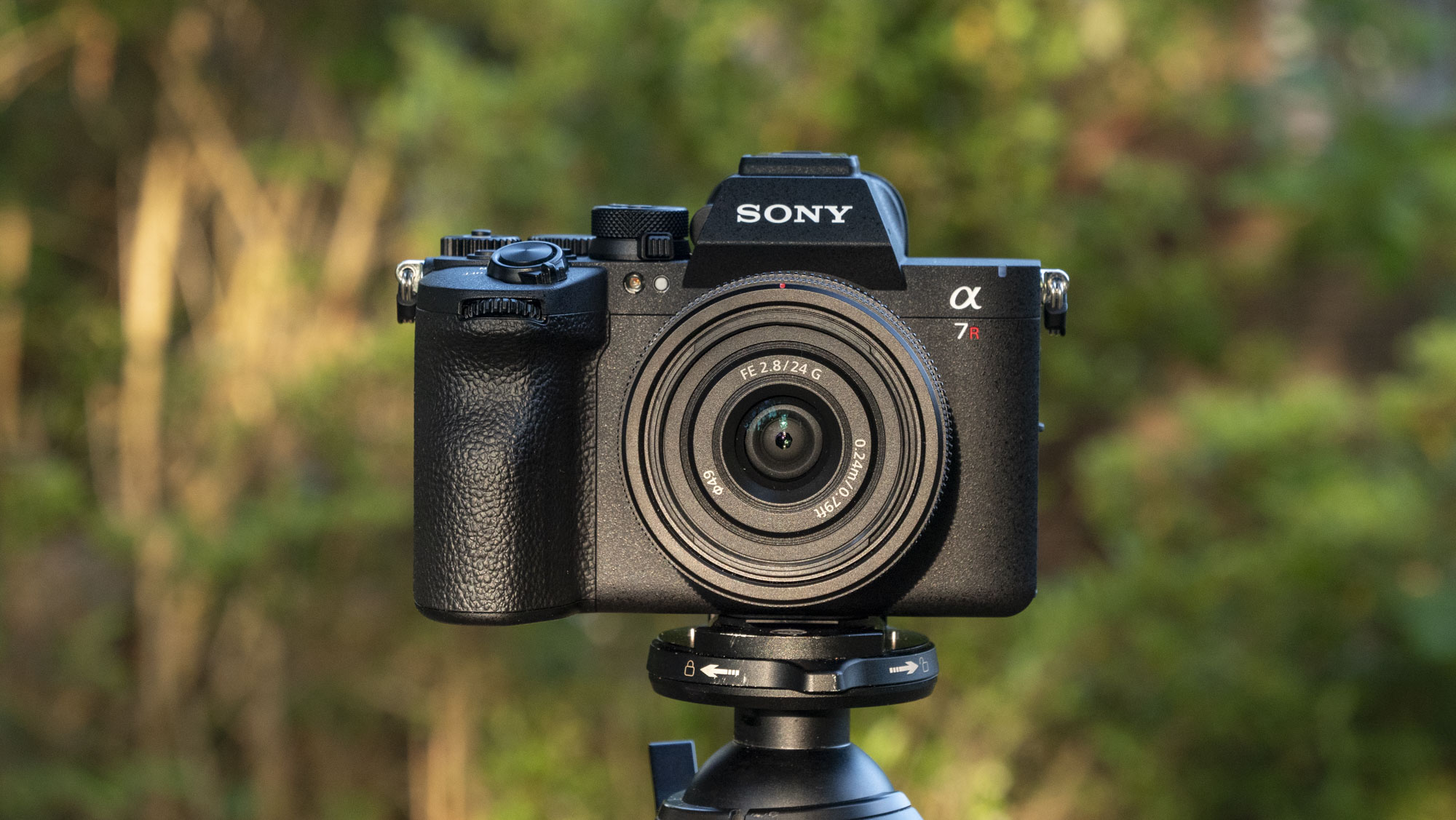 The Sony A7R VI camera with 24mm f/2.8 lens attached, it's mounted to a tripod outdoors, there's a natural background