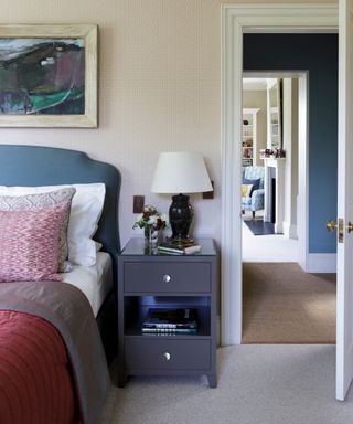 neutral bedroom with subtle wallpapered walls and a navy velvet bed with a navy nightstand and red bed linens