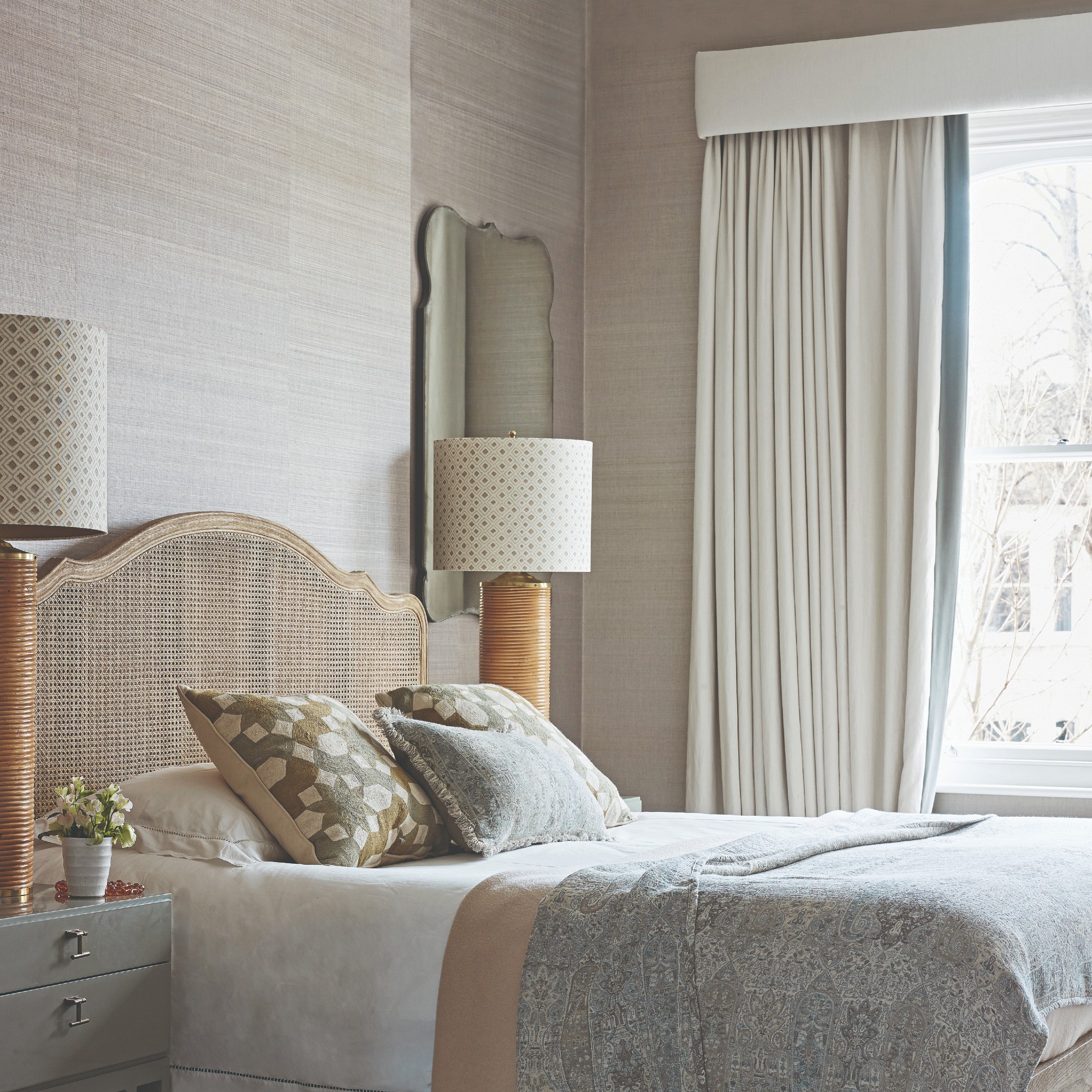 A neutral bedroom with two large table lamps on either side of the bed and window dressed with white heavy curtains and a matching pelmet