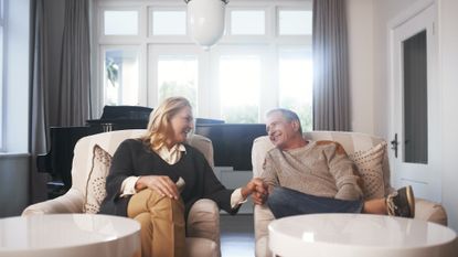 A happy wealthy couple sits in a well-appointed living room, with a grand piano behind them. They are holding hands.