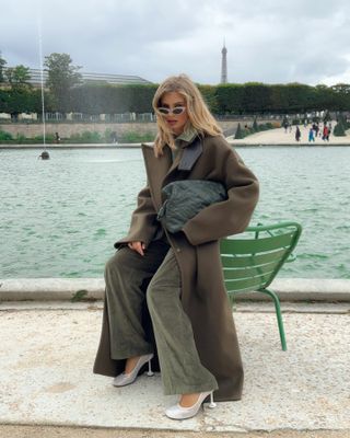 Xenia wears a green wool coat, green corduroy pants, white pumps, and a green clutch bag.