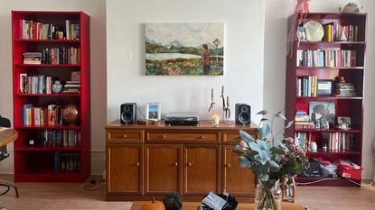 Image of a living room with two red bookcases on either side of a wooden console unit. The console unit has a record player, candlesticks, and a picture frame on it, with a painting hanging overhead.