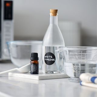 Glass bottle full of white vinegar and a label on the front, next to a pyrex jug and a bottle of essential oils