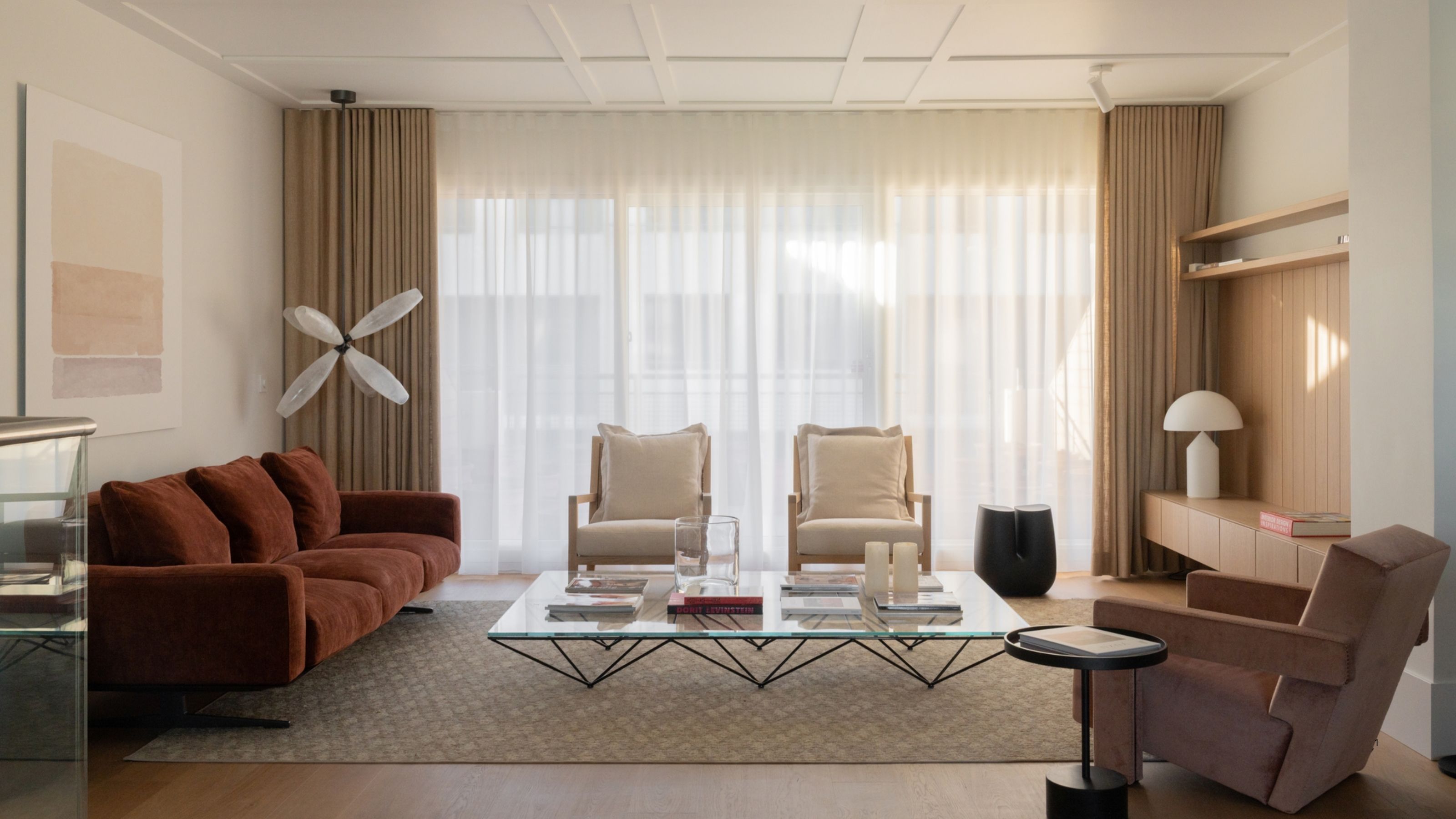A white and beige living room with curtains over the back window wall, a red sofa, white accent chairs, a pink side chair, and a large glass coffee table.