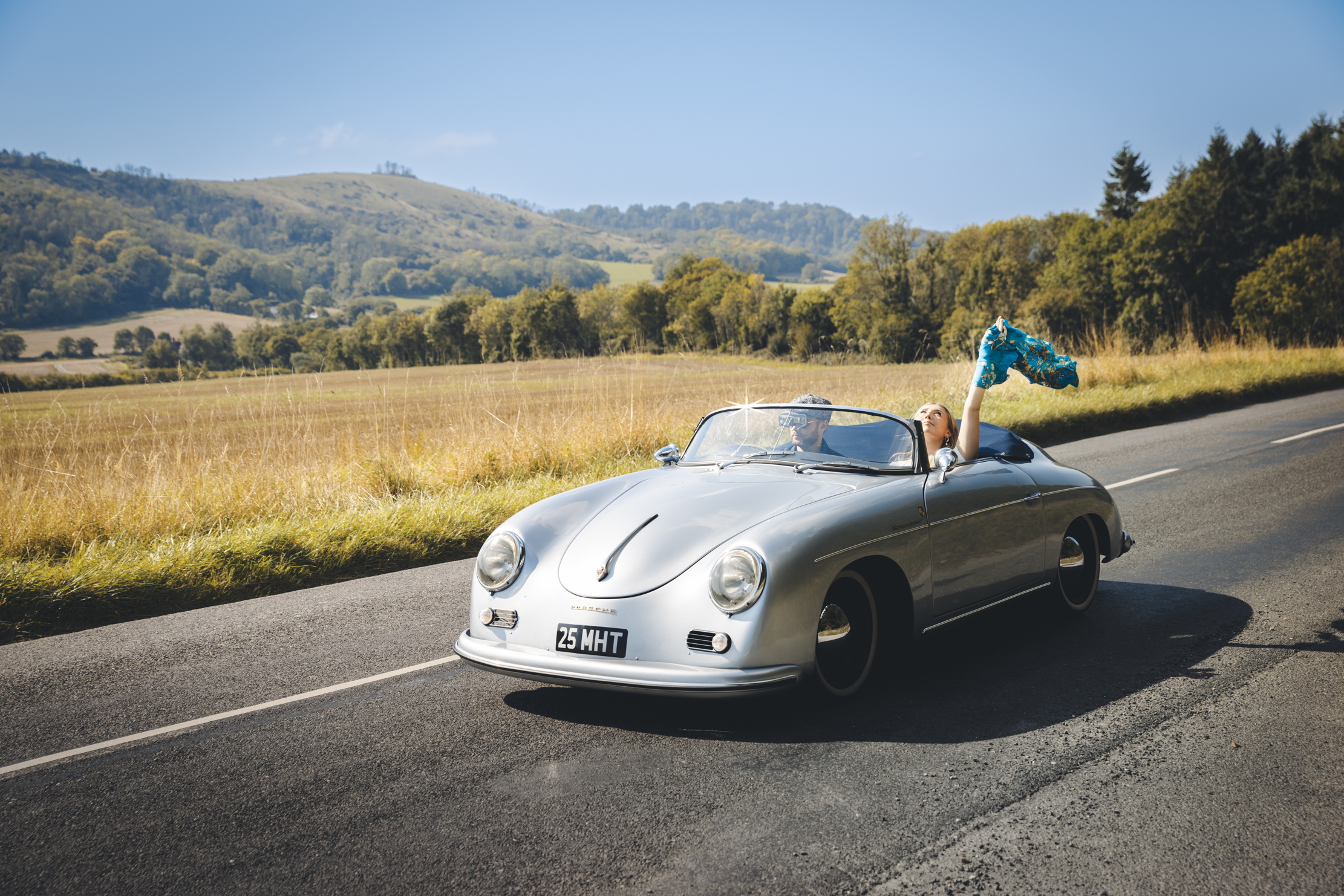 A man and a woman driving through the south downs in a vintage porsche. They look very happy about the whole thing