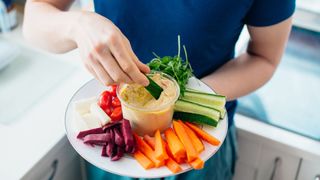 A person holding a plate of assorted cut vegetables around a central dip