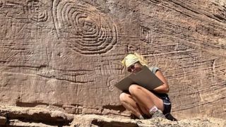 A woman crouches next to petroglyphs. 