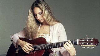 Barbra Streisand wearing blue jeans and a Balinese jacket and playing a guitar.