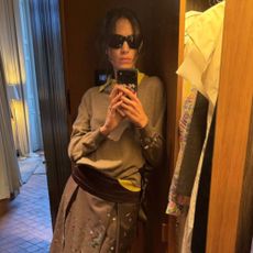 Alexa Chung takes a mirror selfie wearing a knee-length pleated skirt with a grey jumper, a yellow shirt and a purple cummerbund.