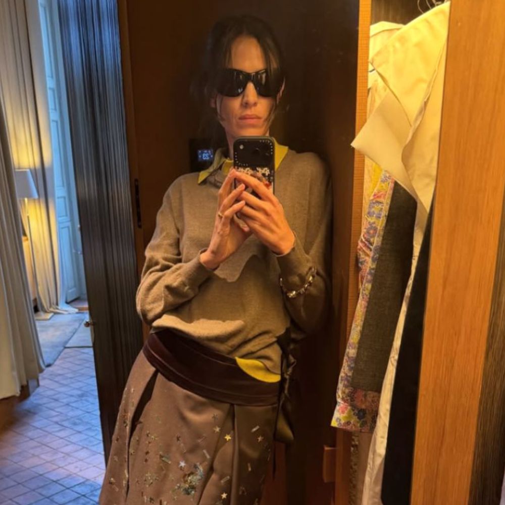 Alexa Chung takes a mirror selfie wearing a knee-length pleated skirt with a grey jumper, a yellow shirt and a purple cummerbund.