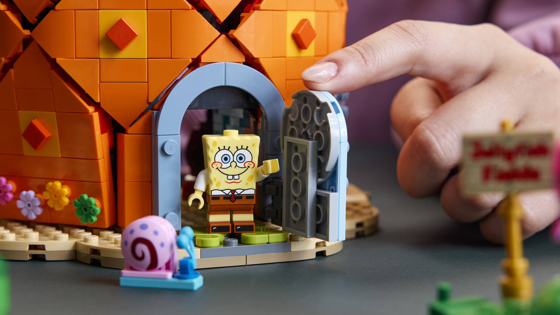 A hand opens the door to Lego Spongebob's house, with a Spongebob minifigure in the doorway waving 