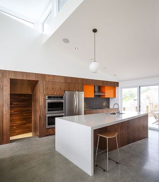 Interior view of the kitchen area at Float House featuring a white sloped ceiling, white walls, grey flooring, spotlights, a sphere pendant light, a wooden and orange kitchen unit with a dark coloured splashback, silver appliances, a wooden and white island with a sink, a wooden stool and glass doors leading to the outside area