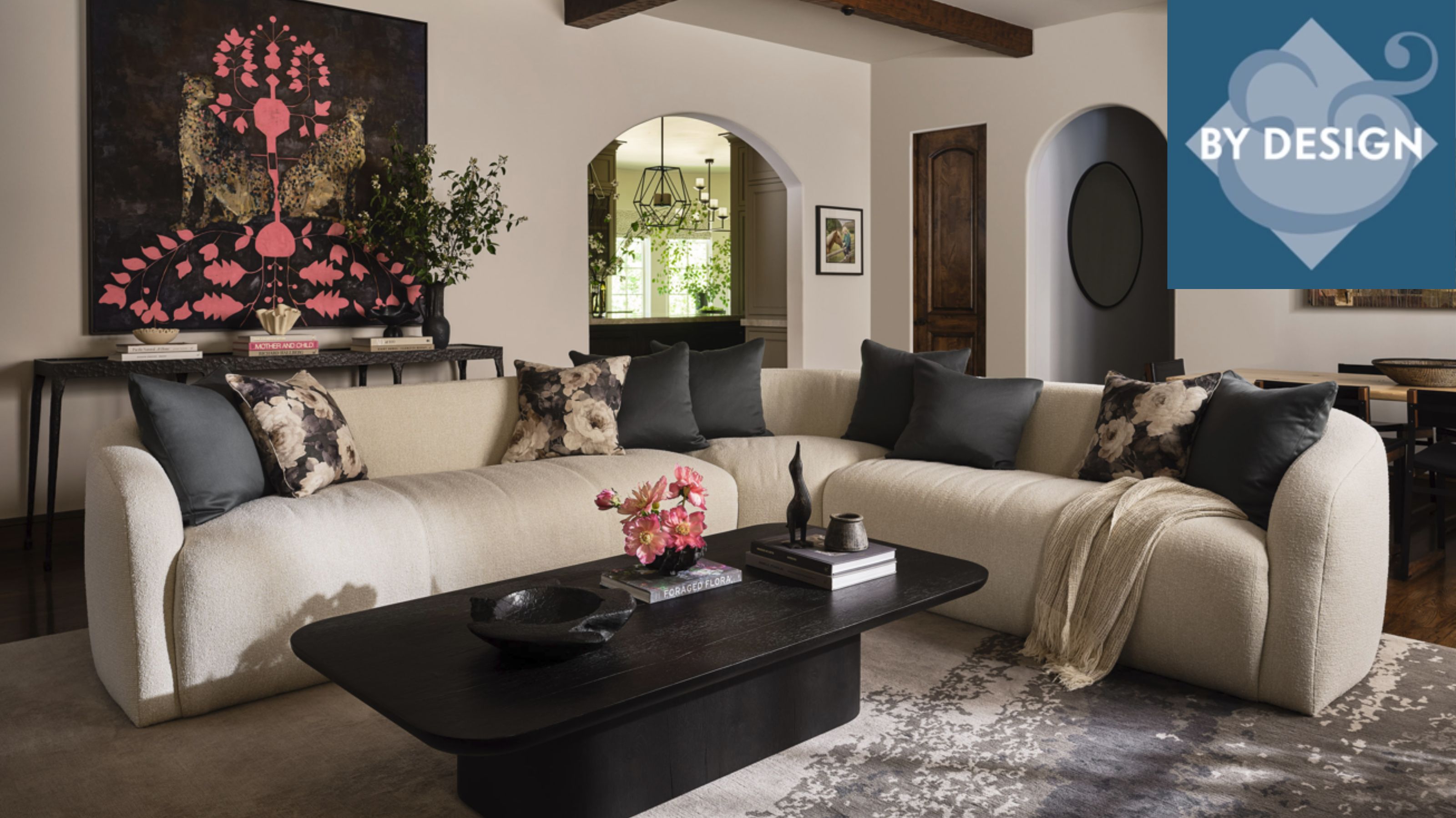 Cozy living room with a large L-Shaped cream sofa with black and floral pillows, an oval-shaped black wood coffee table, a patterned rug, white walls with arch doorways, and wooden beams 